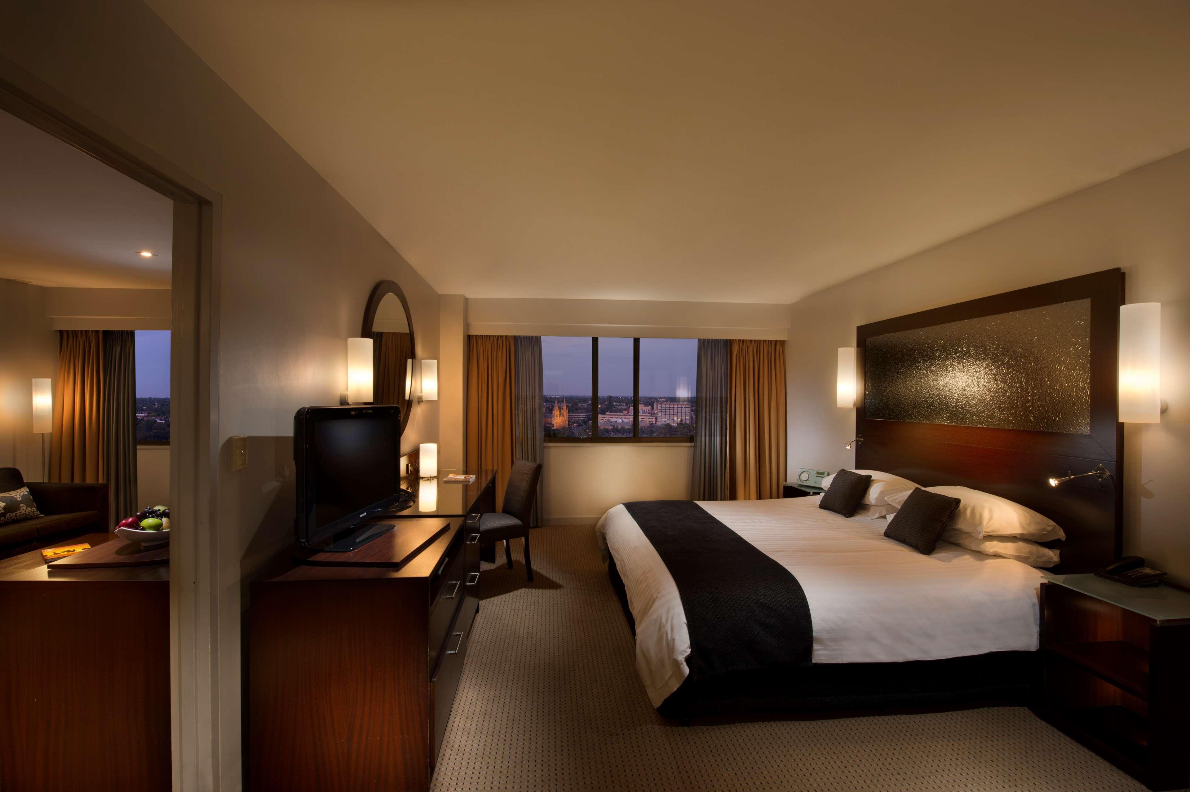 Stamford Plaza Adelaide Hotel Exterior foto The photo features a modern hotel room that includes a large bed with dark and light bedding, complemented by two pillows. The room is well-lit with warm lighting and has a contemporary aesthetic. To one side, there is a wooden dresser with a flat-sc
