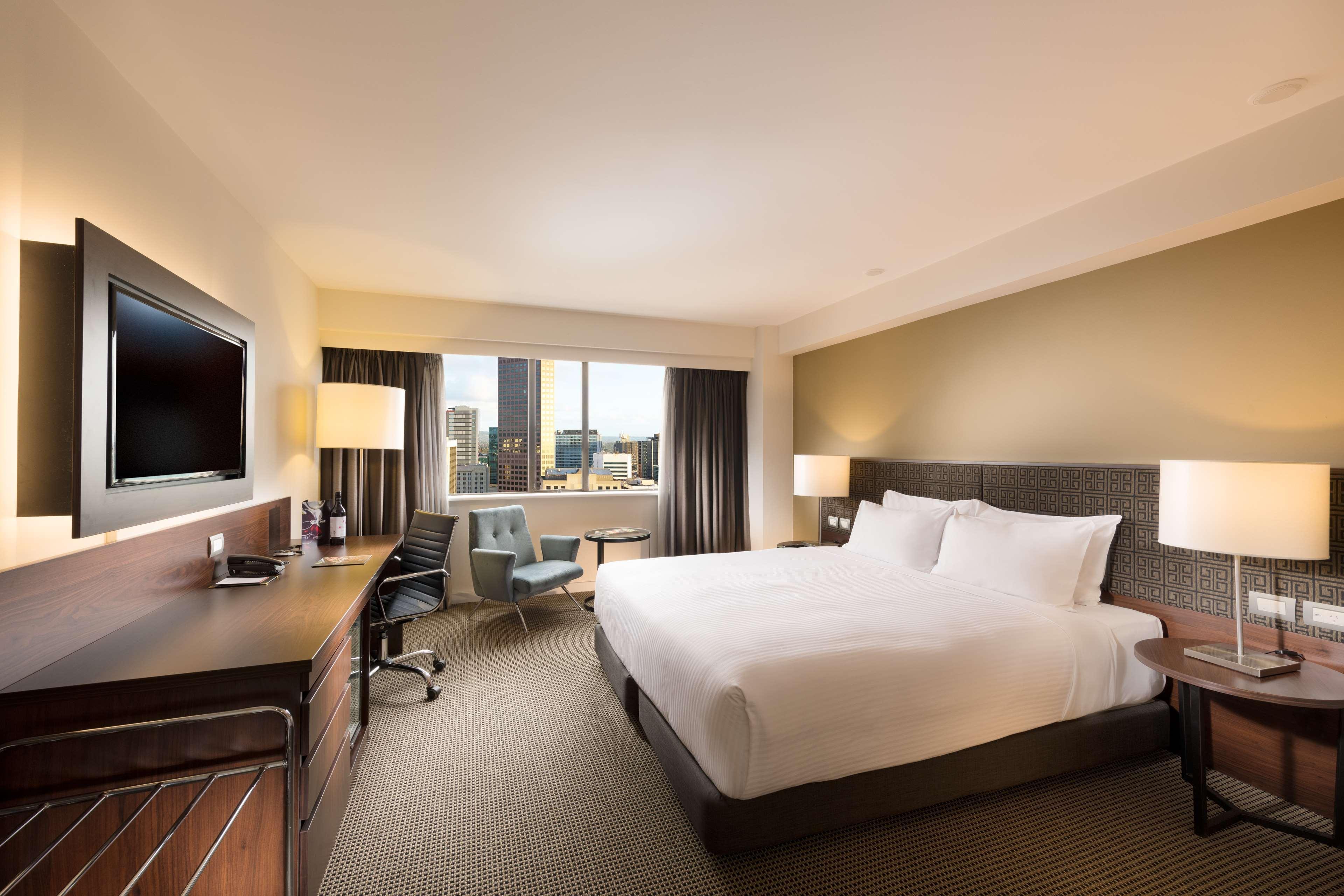 Stamford Plaza Adelaide Hotel Exterior foto The photo shows a modern hotel room with a contemporary design. It features a large bed with white linens, a bedside table on either side with lamps, and a dark wood accent wall. There's a work desk with an office chair and a flat-screen television m